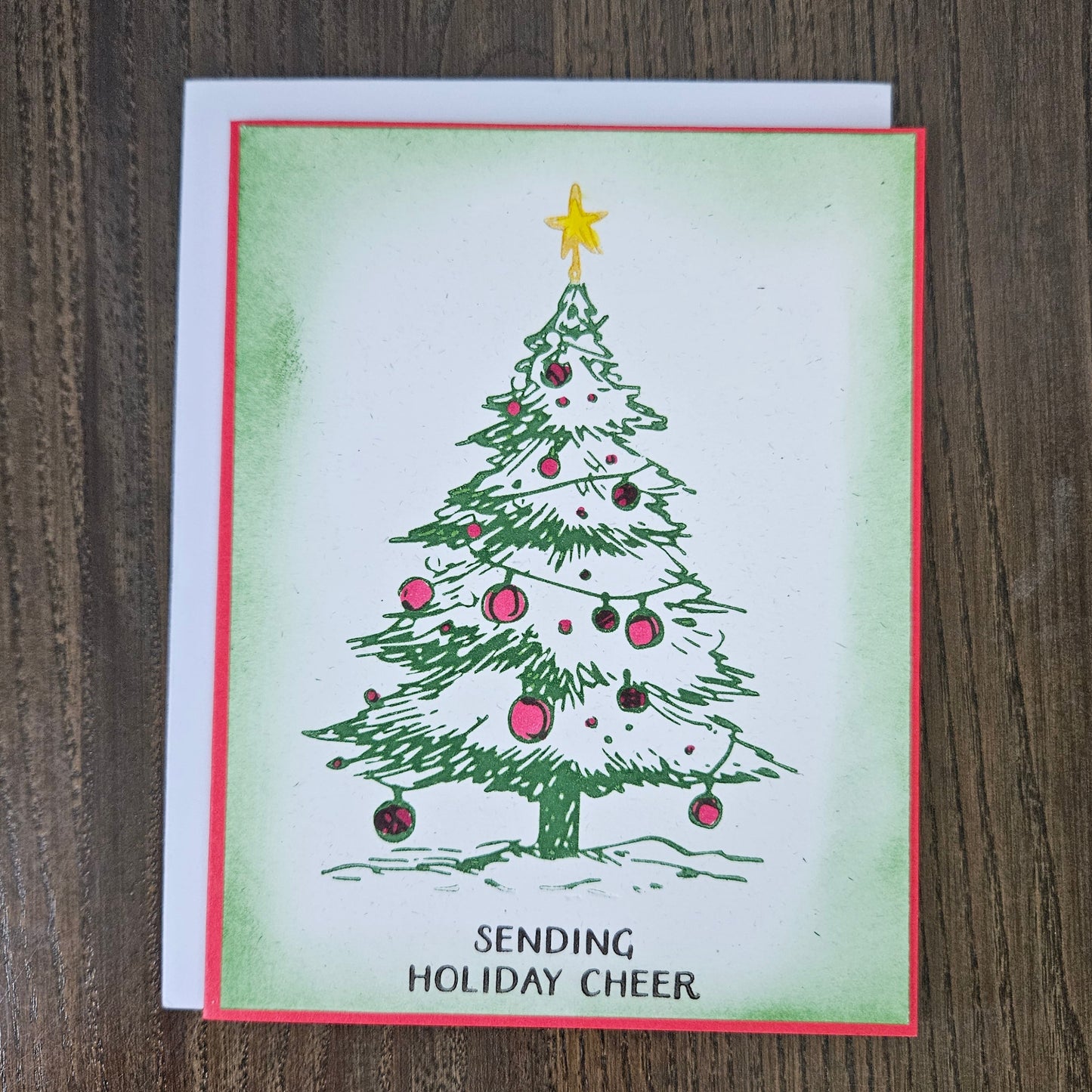 A handmade Christmas greeting card featuring a hand-drawn green Christmas tree with pink and black ornaments, a yellow star, and the text 'SENDING HOLIDAY CHEER' at the bottom. The card is layered over a white backing card and placed on a dark wood background.