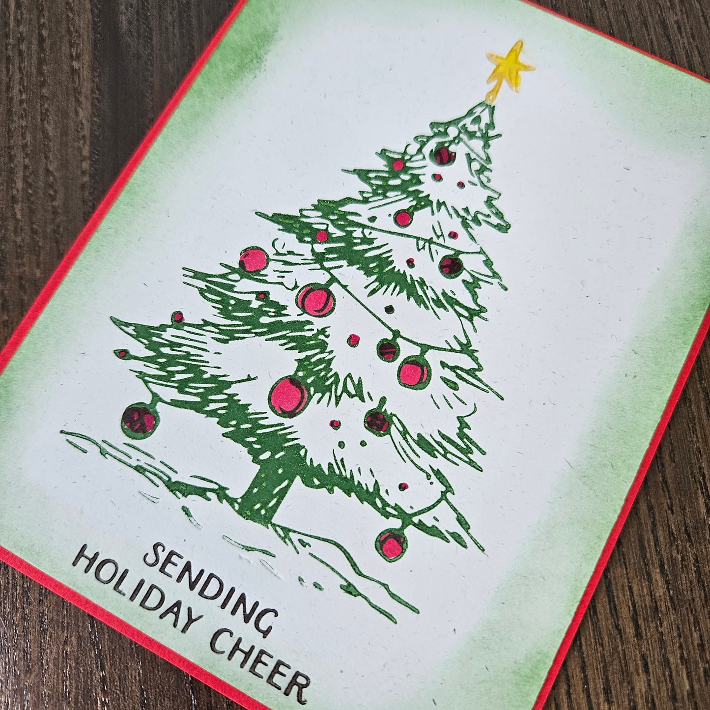 A square greeting card featuring a hand-drawn green Christmas tree with pink ornaments and a yellow star, placed on a dark wood surface. The card has a red border and the text 'SENDING HOLIDAY CHEER' in the lower left corner.