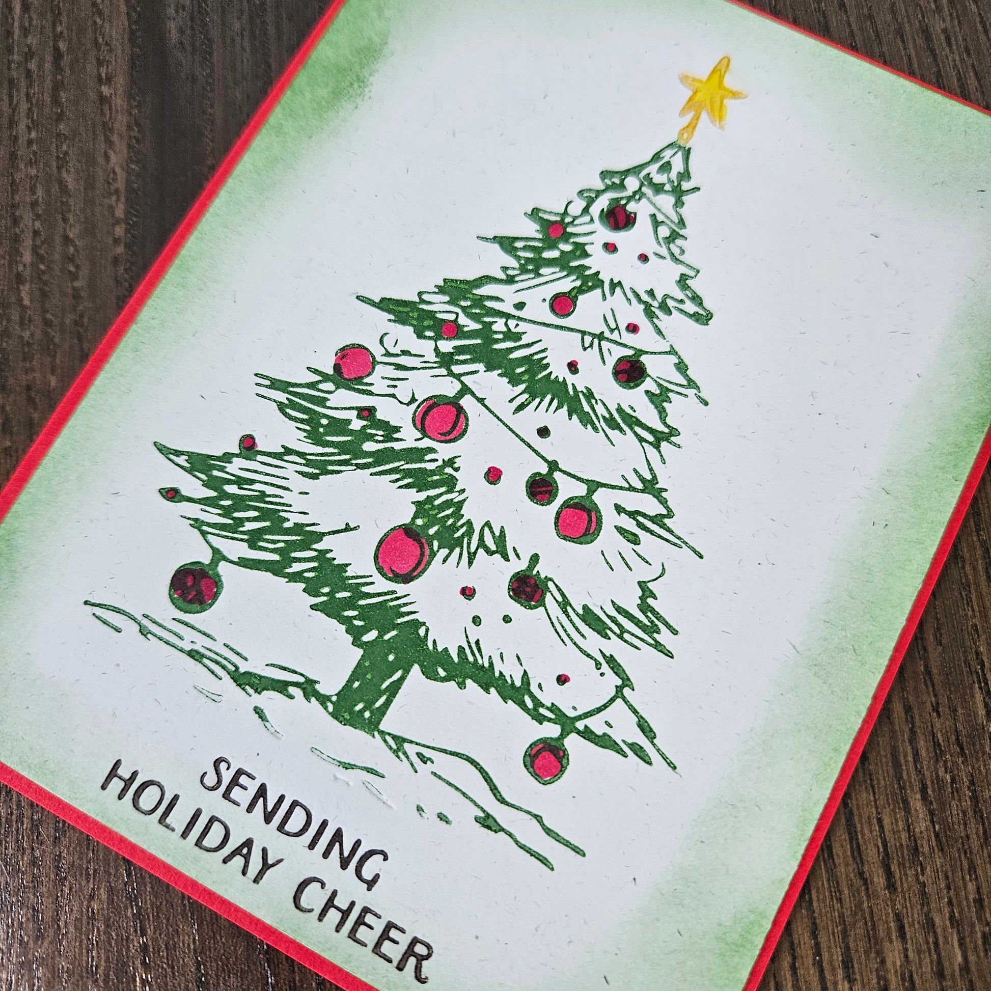 A square greeting card featuring a hand-drawn green Christmas tree with pink ornaments and a yellow star, placed on a dark wood surface. The card has a red border and the text 'SENDING HOLIDAY CHEER' in the lower left corner.