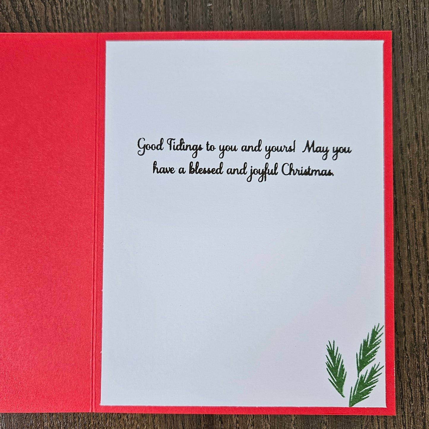 A Christmas greeting card shown open on a dark wood background, with a red outer layer and a white inner card featuring black script text and a small green pine branch illustration in the lower right corner.