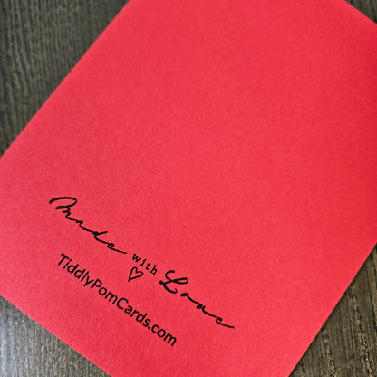 A close-up of a red square card or envelope corner printed with the black script text 'Pamela with Love' and 'TiddlyPomCards.com' on a dark wood surface.