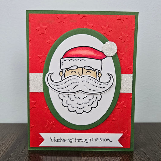 A handmade Christmas greeting card featuring a festive red background with embossed star details, a green border, a white horizontal stripe, and a central oval design of a smiling Santa Claus face with a large white mustache. A white banner at the bottom reads, "stache-ing through the snow.
