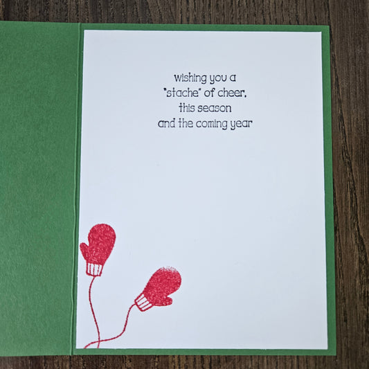 A handmade greeting card with a green border and white center, featuring the printed message 'wishing you a *stache* of cheer, this season and the coming year' above two red glittery mitten illustrations, displayed on a dark wood background.