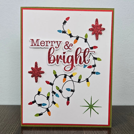 A handmade Christmas greeting card with 'Merry & bright' in red script, decorated with multi-colored string lights, glittery red starbursts, and a green starburst on a white background, standing on a dark wooden surface.