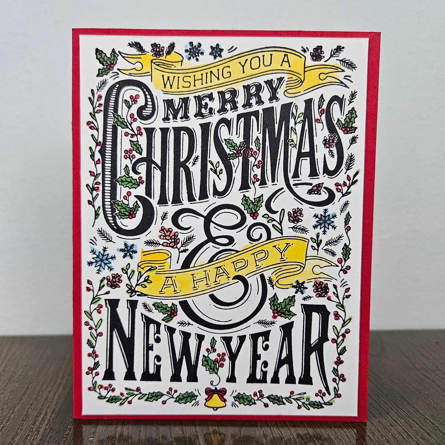 A square Christmas greeting card with a red border, featuring black decorative lettering that reads 'Wishing you a Merry Christmas & Happy New Year' surrounded by holly, pine branches, snowflakes, and yellow ribbon banners, photographed standing on a wooden surface against a light wall.