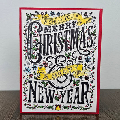 A square Christmas greeting card with a red border, featuring black decorative lettering that reads 'Wishing you a Merry Christmas & Happy New Year' surrounded by holly, pine branches, snowflakes, and yellow ribbon banners, photographed standing on a wooden surface against a light wall.