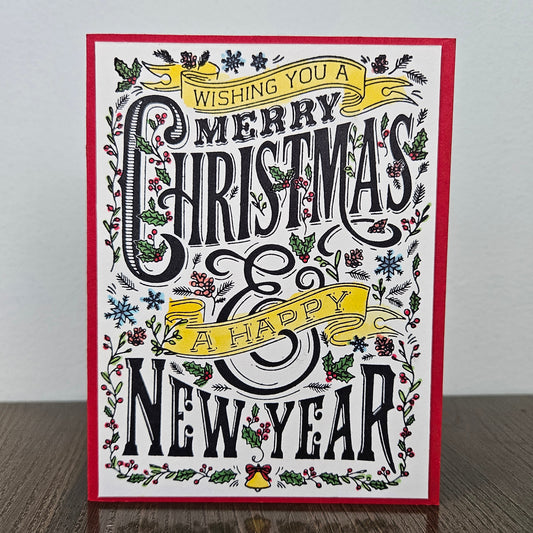 A square Christmas greeting card with a red border, featuring black decorative lettering that reads 'Wishing you a Merry Christmas & Happy New Year' surrounded by holly, pine branches, snowflakes, and yellow ribbon banners, photographed standing on a wooden surface against a light wall.
