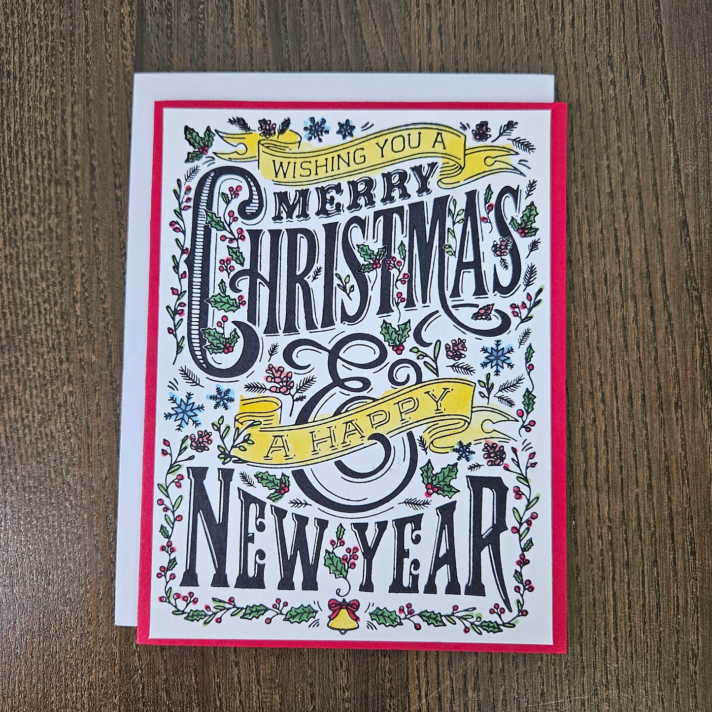 A square Christmas greeting card with festive hand-lettered text reading 'Wishing you a Merry Christmas & Happy New Year,' surrounded by illustrated holly, mistletoe, snowflakes, and small bells. The card is photographed on a brown wood surface, with a white cardstock backing visible behind it.