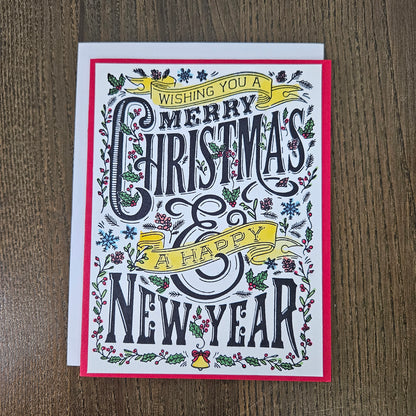 A square Christmas greeting card with festive hand-lettered text reading 'Wishing you a Merry Christmas & Happy New Year,' surrounded by illustrated holly, mistletoe, snowflakes, and small bells. The card is photographed on a brown wood surface, with a white cardstock backing visible behind it.