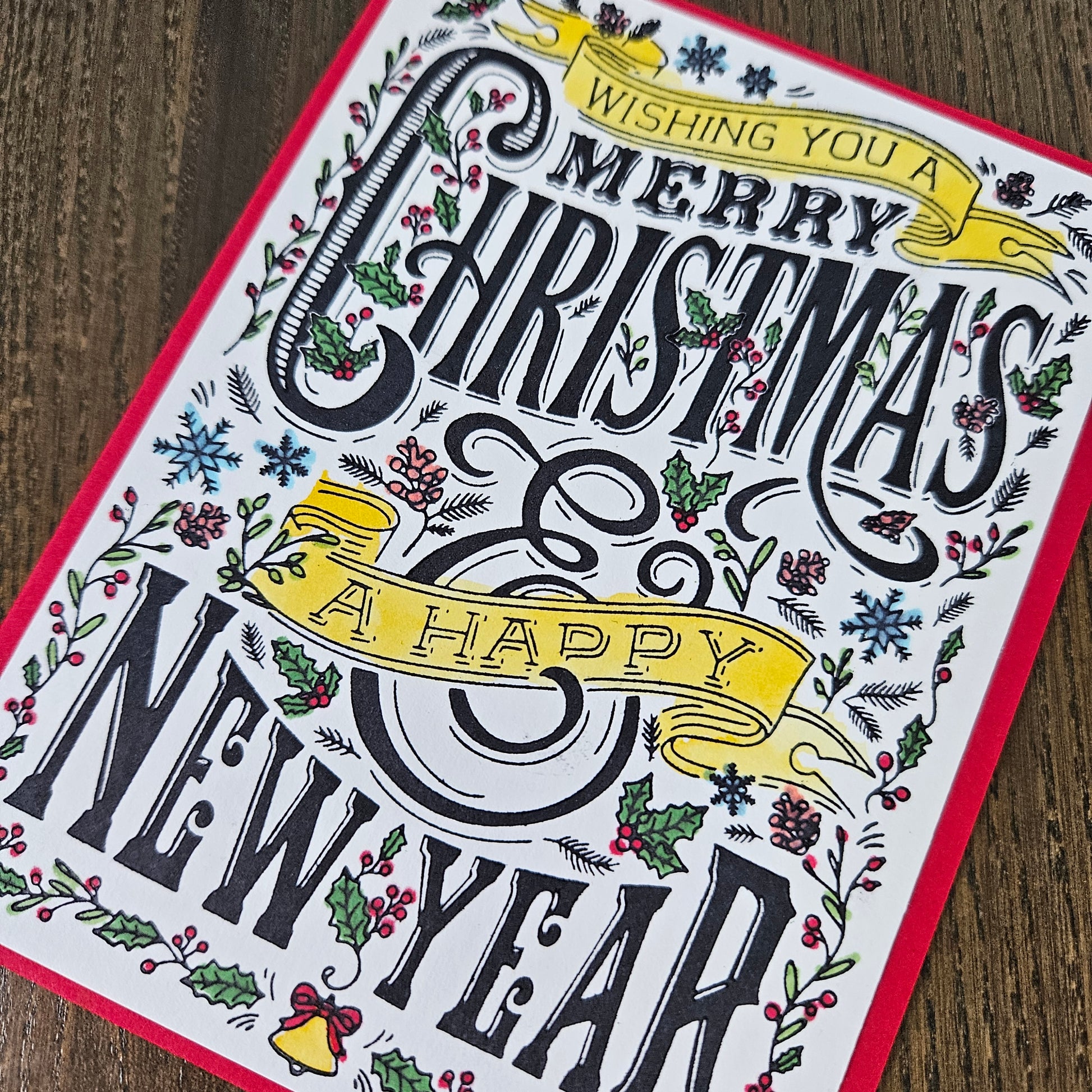 A square greeting card featuring festive black typography that reads 'Wishing you a Merry Christmas & Happy New Year' surrounded by illustrated holly, berries, snowflakes, and yellow ribbon banners, photographed on a wooden surface.