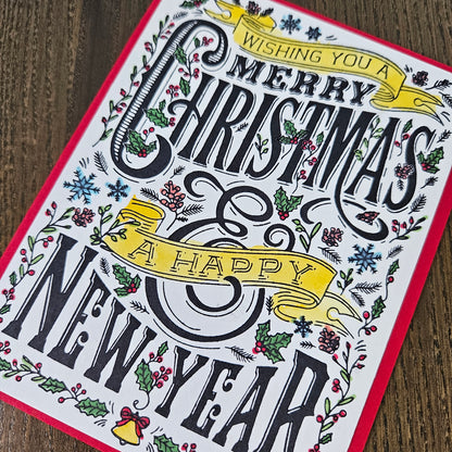 A square greeting card featuring festive black typography that reads 'Wishing you a Merry Christmas & Happy New Year' surrounded by illustrated holly, berries, snowflakes, and yellow ribbon banners, photographed on a wooden surface.