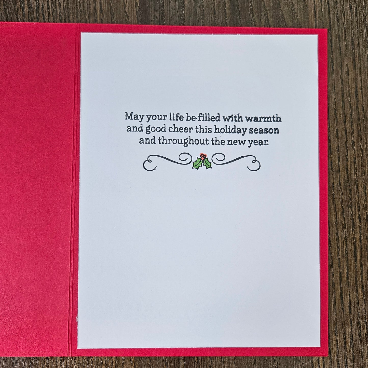 A square holiday greeting card with a bright red envelope, shown open on a dark wood background. The white card features a black printed holiday message with a small holly graphic.