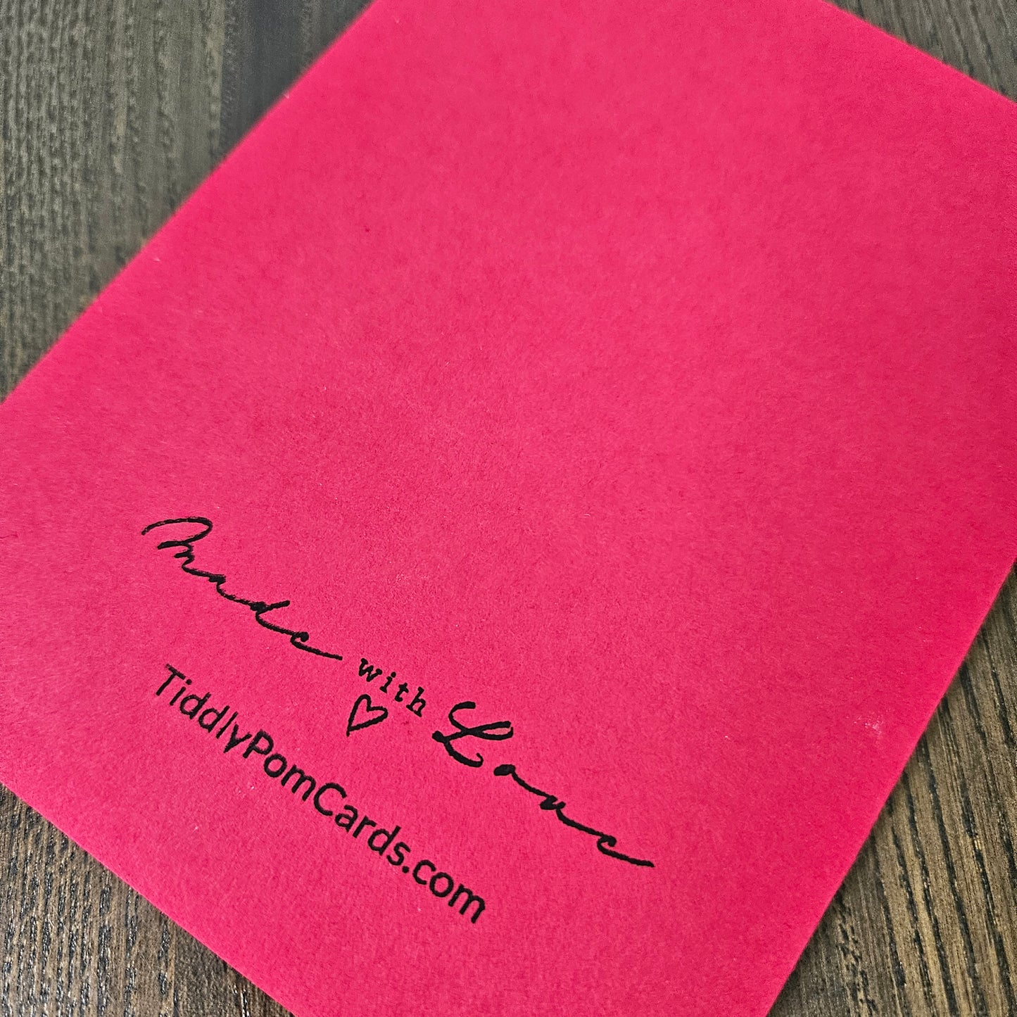 A bright pink square card or paper sheet placed on a dark wood surface, printed with the black script text 'Made with Love' and 'TiddlyPomCards.com' near the lower left.