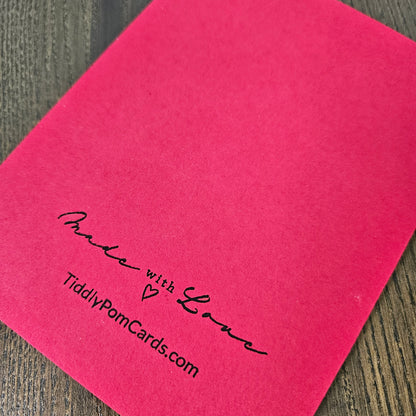 A bright pink square card or paper sheet placed on a dark wood surface, printed with the black script text 'Made with Love' and 'TiddlyPomCards.com' near the lower left.
