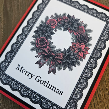 A close-up of a handmade Christmas greeting card featuring a black and red floral wreath with the text 'Merry Gothmas' on a white background, bordered by a black lace pattern and red cardstock edges, resting on a wooden surface.