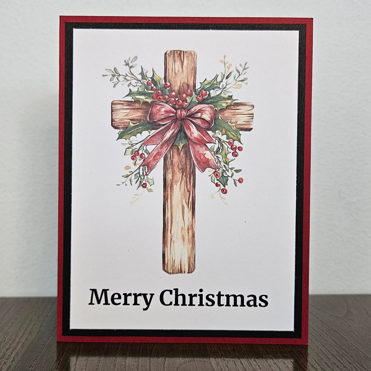 A square Christmas greeting card with a red and black border, featuring a watercolor-style wooden cross decorated with greenery and a pink bow, with the text 'Merry Christmas' at the bottom, displayed on a dark wooden surface against a light wall.