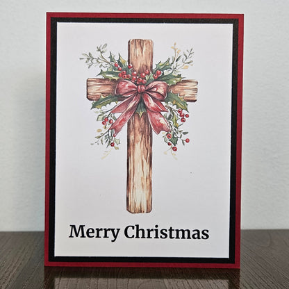 A square Christmas greeting card with a red and black border, featuring a watercolor-style wooden cross decorated with greenery and a pink bow, with the text 'Merry Christmas' at the bottom, displayed on a dark wooden surface against a light wall.