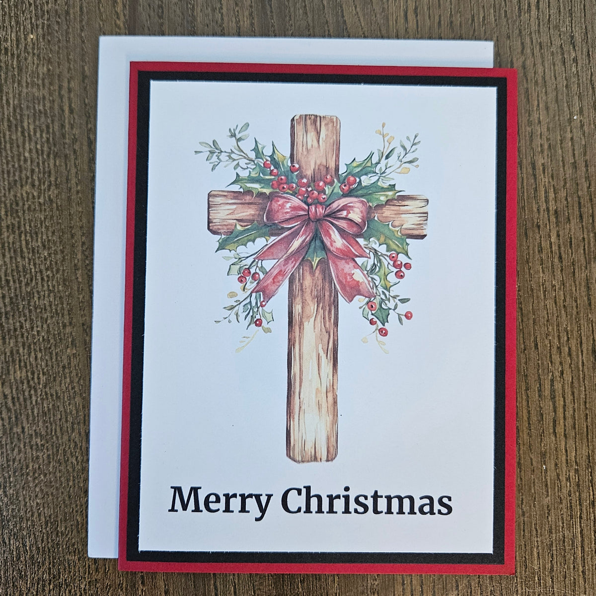 A Christmas greeting card with a wooden cross decorated with green holly leaves and red berries, tied with a pink bow. The card reads 'Merry Christmas' in black text at the bottom.