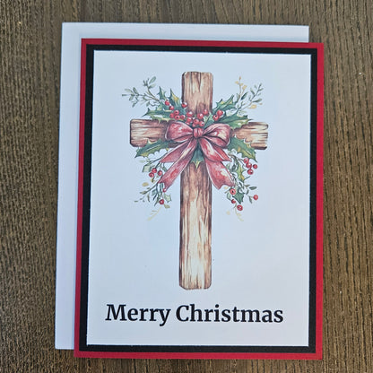 A Christmas greeting card with a wooden cross decorated with green holly leaves and red berries, tied with a pink bow. The card reads 'Merry Christmas' in black text at the bottom.