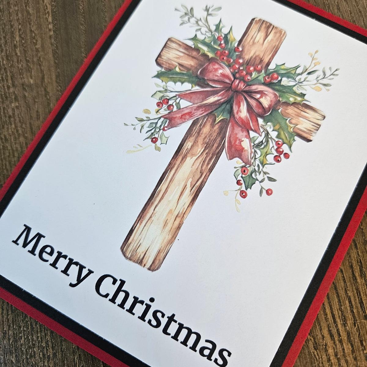 A square Christmas greeting card featuring a rustic wooden cross decorated with holly leaves, red berries, and a pinkish-red bow, with the text 'Merry Christmas' printed at the bottom. The card is photographed on a wooden surface.