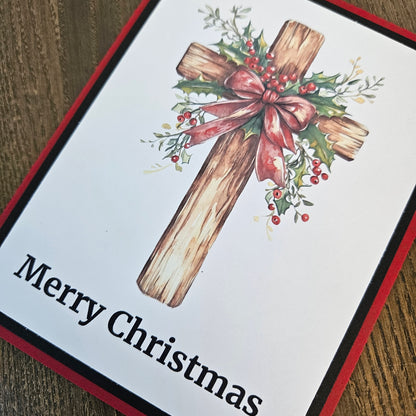 A square Christmas greeting card featuring a rustic wooden cross decorated with holly leaves, red berries, and a pinkish-red bow, with the text 'Merry Christmas' printed at the bottom. The card is photographed on a wooden surface.
