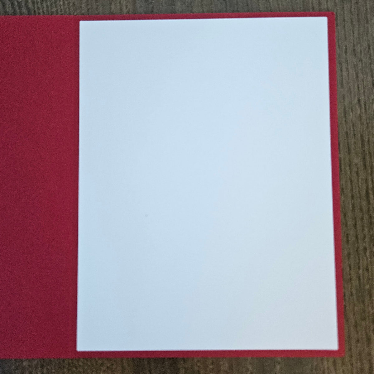 A blank white rectangular card or paper insert placed on top of a textured red sheet, photographed from above on a brown wood-grain surface.