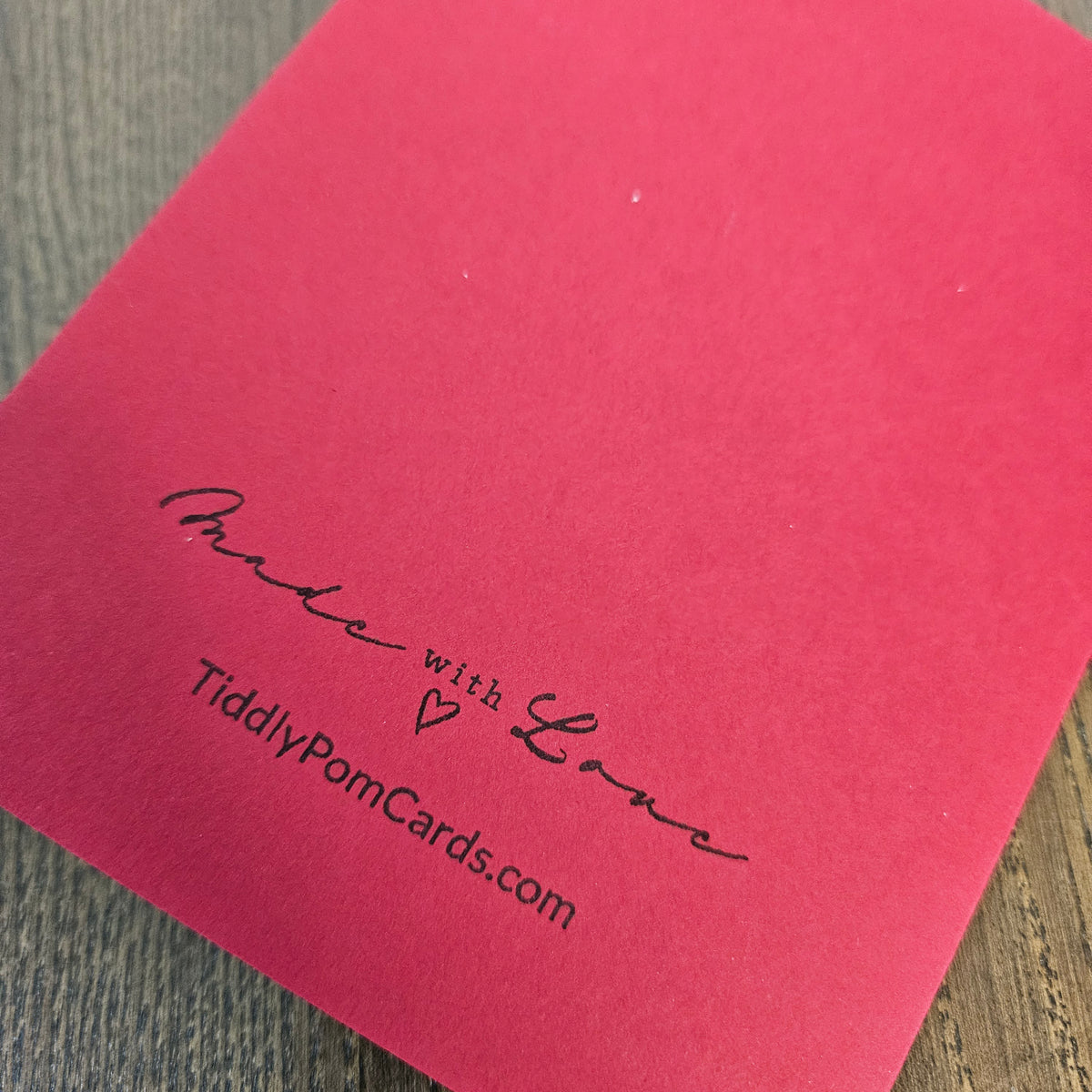 A close-up of a bright pink square card with black printed text reading 'Made with Love' and 'TiddlyPomCards.com' on a wood surface.