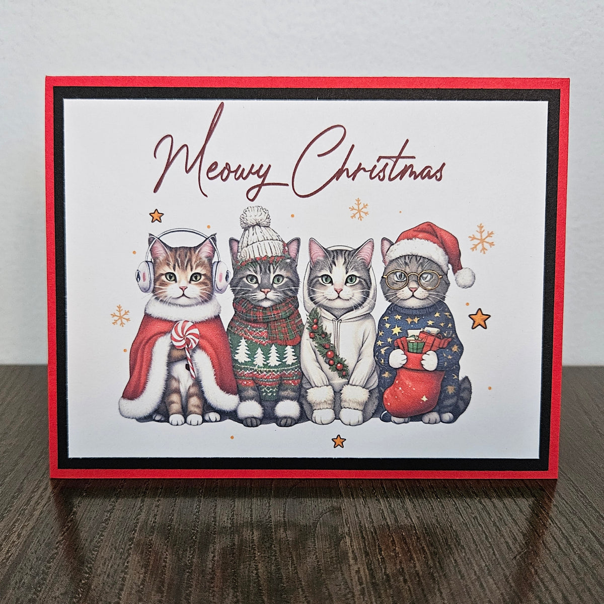 A square Christmas greeting card featuring four illustrated cats dressed in holiday clothing, with the words 'Mewzy Christmas' at the top. The card has a red border and is photographed standing on a dark wooden surface against a light wall.