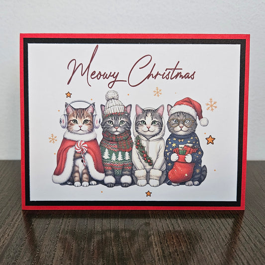 A square Christmas greeting card featuring four illustrated cats dressed in holiday clothing, with the words 'Mewzy Christmas' at the top. The card has a red border and is photographed standing on a dark wooden surface against a light wall.