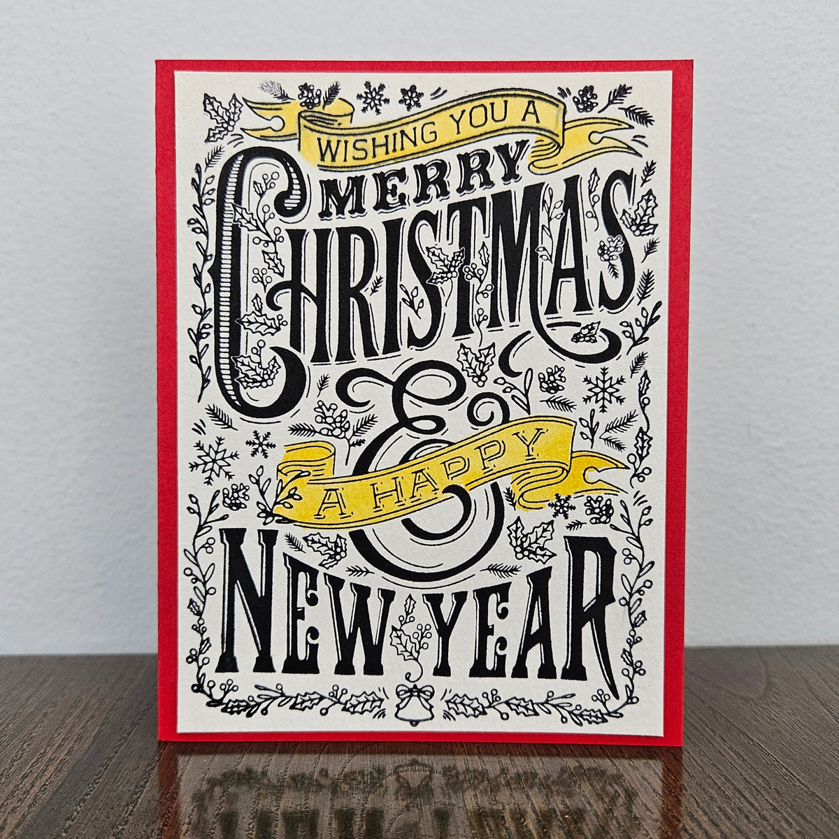 A square Christmas greeting card with a red border, featuring black and white decorative typography that reads 'Wishing you a Merry Christmas & Happy New Year,' styled with floral illustrations and yellow ribbon banners, standing on a dark wooden surface against a light gray wall.