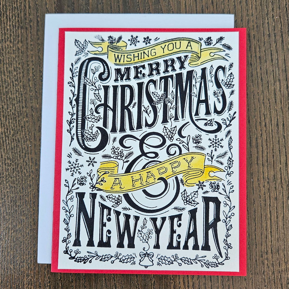 A festive greeting card with a red border and white background, featuring black decorative lettering that reads 'Wishing you a Merry Christmas & A Happy New Year' with yellow ribbon banners, shown on a wooden surface with a white envelope behind it.