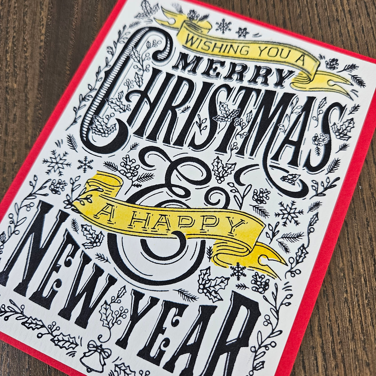 A square greeting card with a red border, featuring black and yellow typography that reads 'Wishing You a Merry Christmas & A Happy New Year' surrounded by delicate holiday illustrations, shown on a dark wood background.