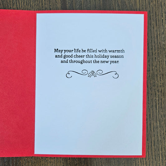 An open red greeting card lying on a dark wood surface, showing a white insert with black holiday text and a small decorative flourish.