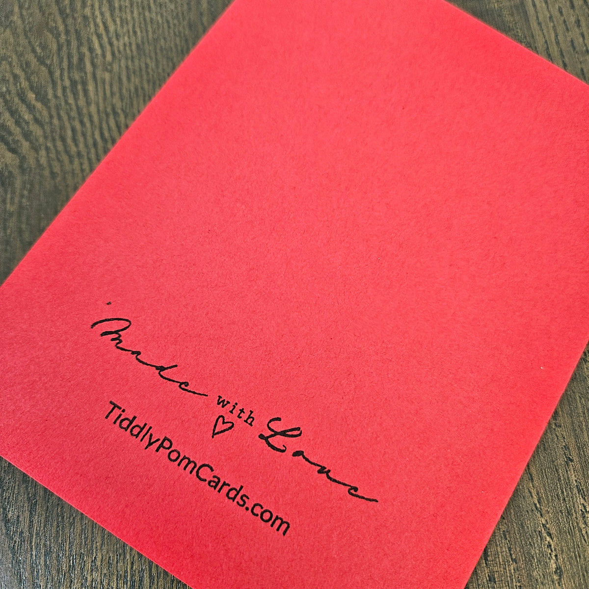 A red square card or envelope cover photographed at an angle on a dark wood surface, with black printed text reading “Made with Love” and “TiddlyPomCards.com” near the lower left.
