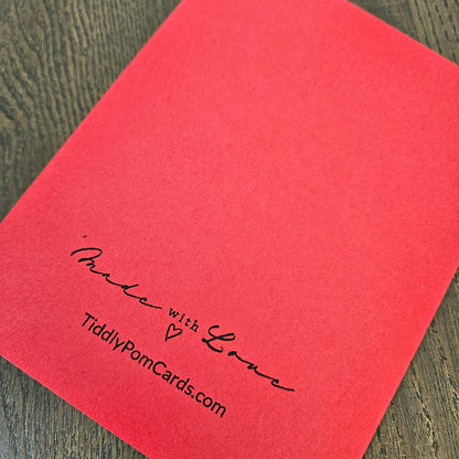 A red square card or envelope cover photographed at an angle on a dark wood surface, with black printed text reading “Made with Love” and “TiddlyPomCards.com” near the lower left.