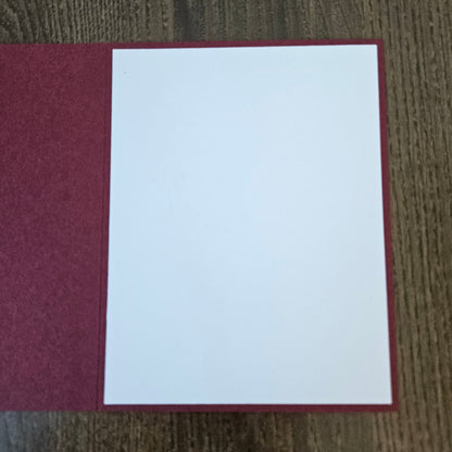 A blank white card or paper insert placed on top of a textured dark red folder or book cover, photographed from above on a wood-grain surface.