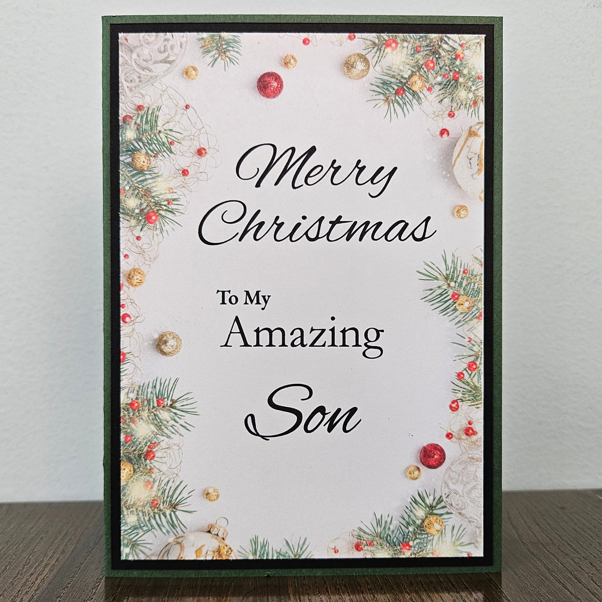 A personalized Christmas greeting card with a dark green border, featuring holiday greenery, red berries, and gold ornaments around the border. The center has a white background with black script text reading 'Merry Christmas To My Amazing Son'.