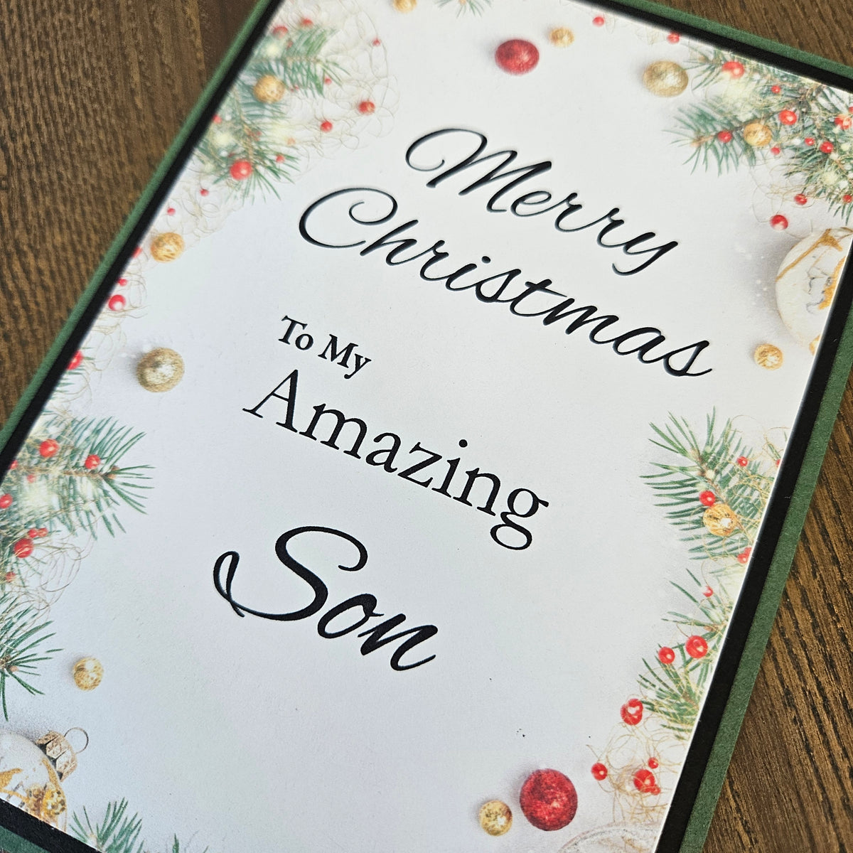 A personalized Christmas greeting card with the text 'Merry Christmas To My Amazing Son' in black script and serif fonts, decorated with green pine branches, red and gold ornaments, and a dark green border, shown on a wooden surface.