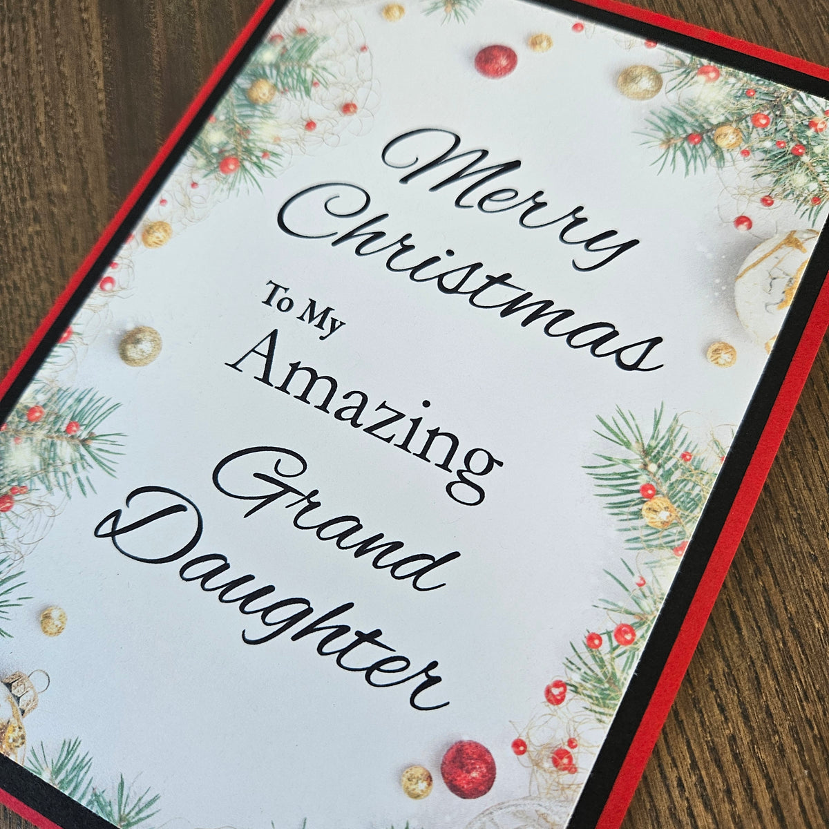 A square Christmas greeting card with the text 'Merry Christmas To My Amazing Granddaughter' in black script, decorated with green pine branches, red and gold ornaments, and a red border, shown on a wooden surface.