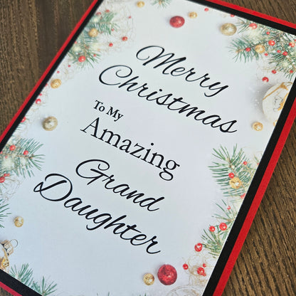 A square Christmas greeting card with the text 'Merry Christmas To My Amazing Granddaughter' in black script, decorated with green pine branches, red and gold ornaments, and a red border, shown on a wooden surface.
