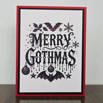 A handmade gothic-themed greeting card standing upright on a dark wooden surface against a light gray wall. The card features a white background with a red and black double border, decorated with black gothic lettering that reads 'MERRY GOTHMAS', bats, holly, snowflakes, and ornaments.
