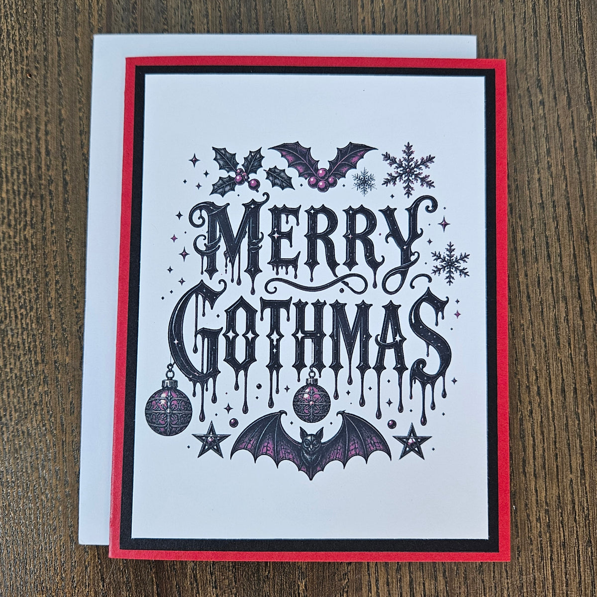 A square Christmas greeting card with a white background, featuring the text 'MERRY GOTHMAS' in a black gothic font. The card is decorated with gothic illustrations including bats, holly, snowflakes, stars, and ornaments in black and purple.