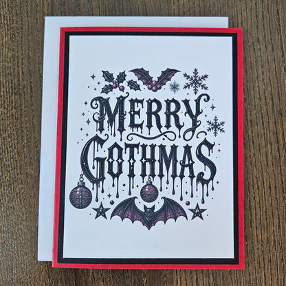A square Christmas greeting card with a white background, featuring the text 'MERRY GOTHMAS' in a black gothic font. The card is decorated with gothic illustrations including bats, holly, snowflakes, stars, and ornaments in black and purple.