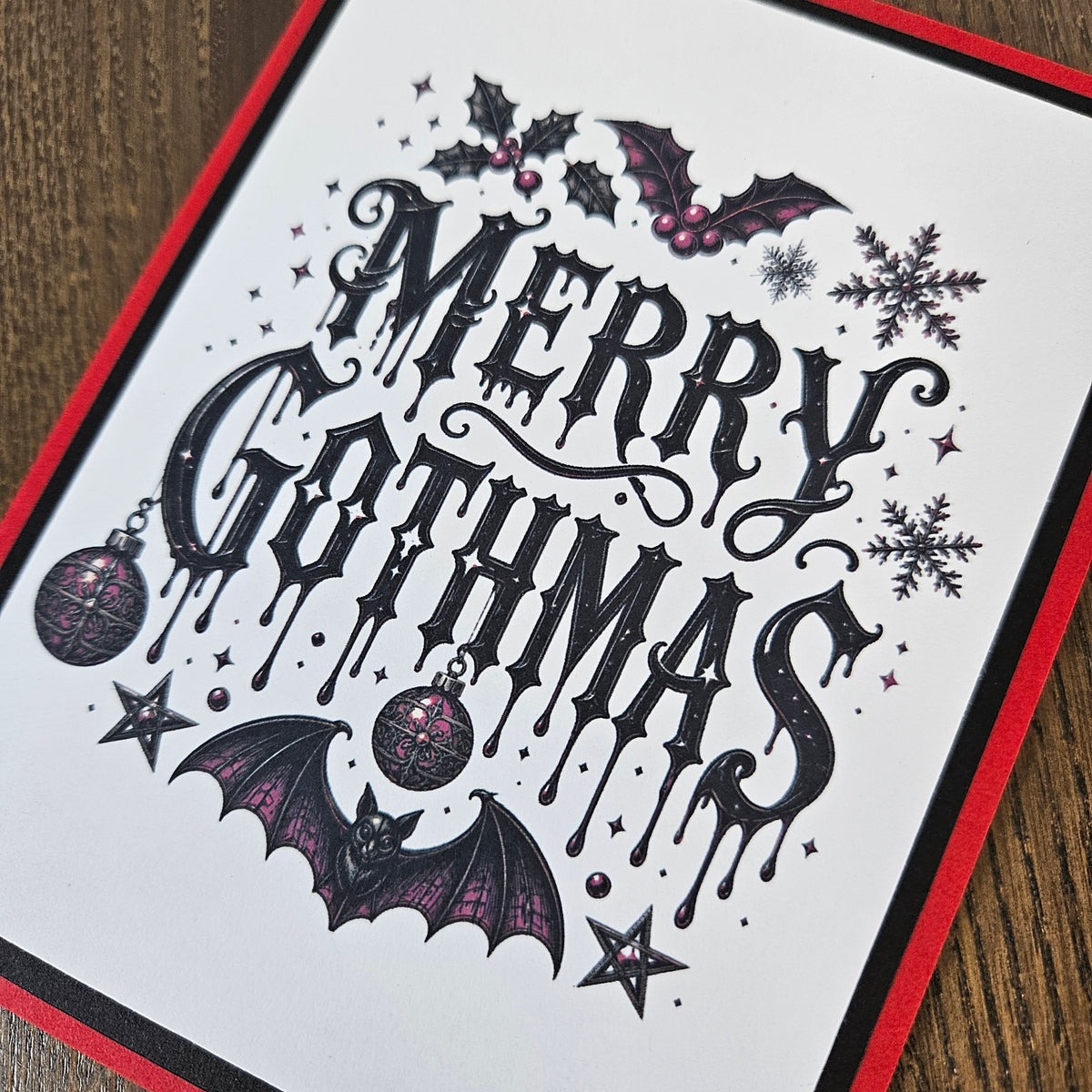 A close-up of a white greeting card featuring gothic-style 'Merry Gothas' lettering with bats, holly, snowflakes, stars, and ornaments, bordered in red and black, photographed on a wooden surface.