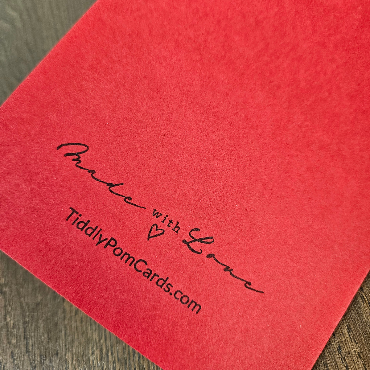 A close-up of a red textured card or paper with black printed text reading 'Made with Love' and 'TiddlyPomCards.com' on a wood surface.