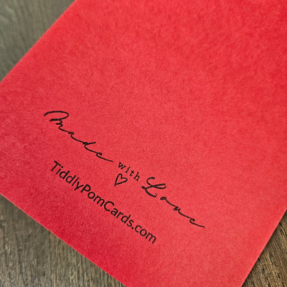 A close-up of a red textured card or paper with black printed text reading 'Made with Love' and 'TiddlyPomCards.com' on a wood surface.