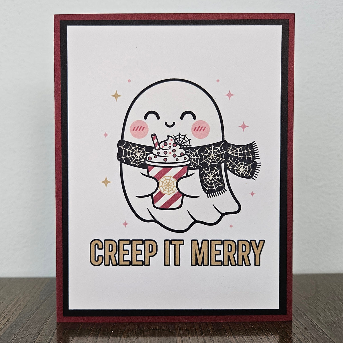 A handmade greeting card featuring a cute cartoon ghost holding a hot cocoa drink, with the text 'CREEP IT MERRY' below. The card has a dark red border, a black inner border, and is displayed standing on a dark wooden surface against a light gray wall.