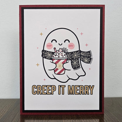 A handmade greeting card featuring a cute cartoon ghost holding a hot cocoa drink, with the text 'CREEP IT MERRY' below. The card has a dark red border, a black inner border, and is displayed standing on a dark wooden surface against a light gray wall.