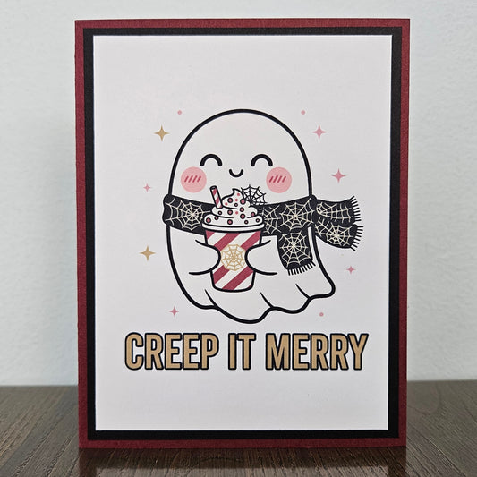A handmade greeting card featuring a cute cartoon ghost holding a hot cocoa drink, with the text 'CREEP IT MERRY' below. The card has a dark red border, a black inner border, and is displayed standing on a dark wooden surface against a light gray wall.