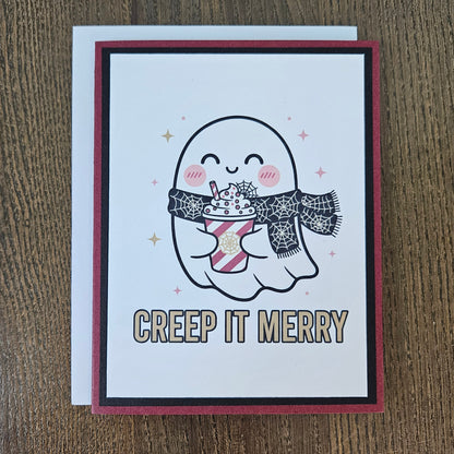 A handmade greeting card featuring a cute cartoon ghost holding a hot drink, with the text 'CREEP IT MERRY' below, displayed on a wooden tabletop next to its white envelope.